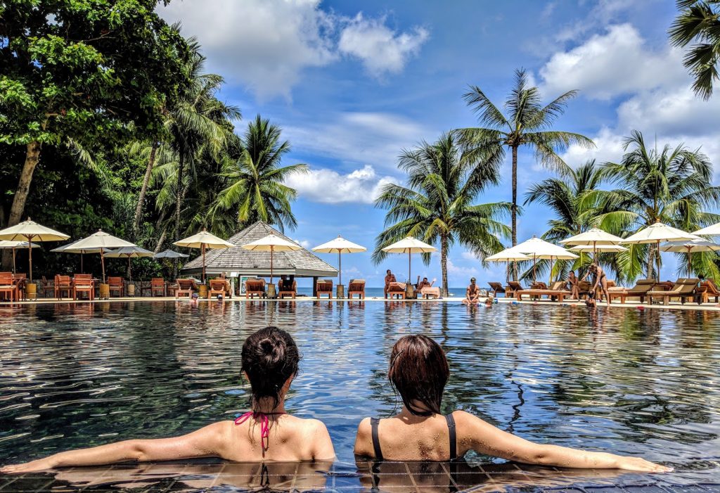 duas mulheres de costas dentro de piscina de hotel cercada de mesas e cadeiras de frente para o mar