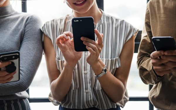 Três pessoas usam seus celulares. A imagem mostra apenas troncos e mãos, exceto a mulher no centro, que mostra o sorriso.