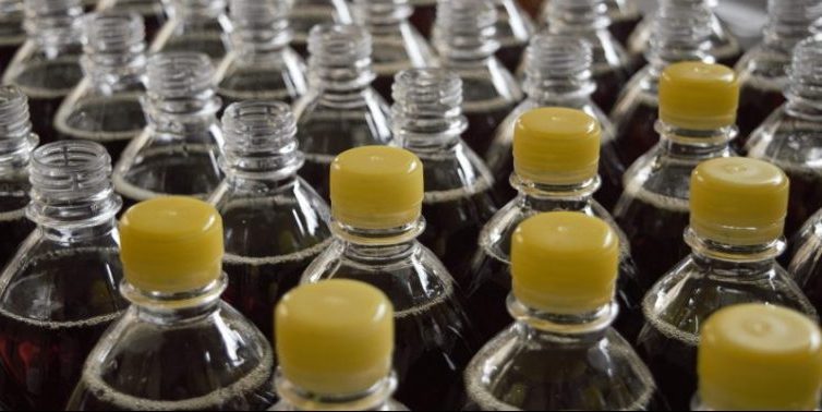 Diversas garrafas de corpo transparente enfileiradas em linha de produção. Algumas possuem tampas amarelas.