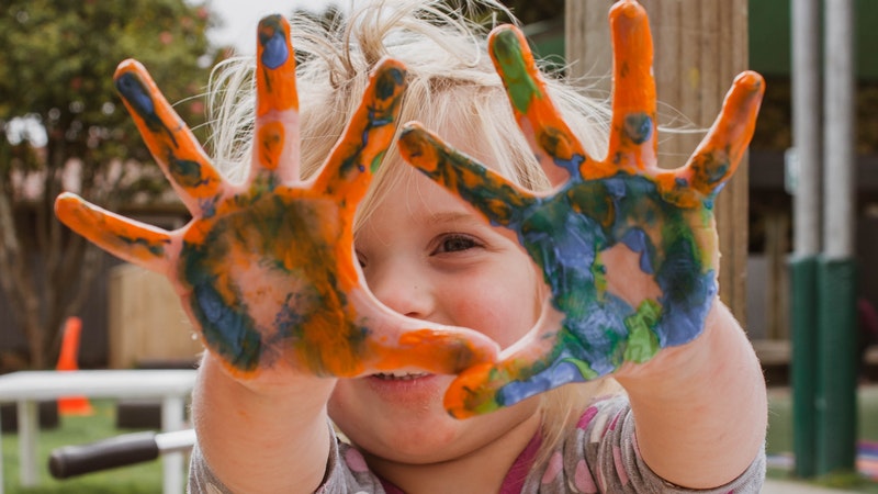 Criança pequena mostra as mãos espalmadas cheias de tintas coloridas.