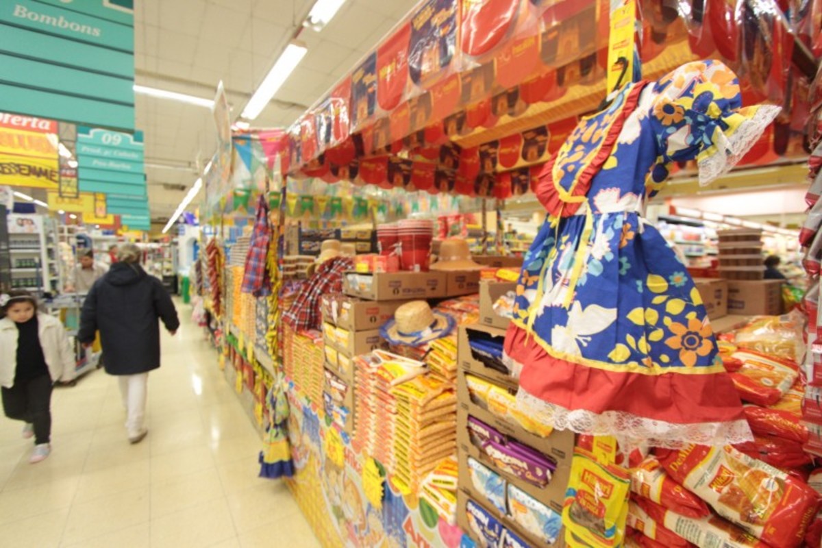 Em foco, ilha temática de supermercado com itens de festa junina. No fundo, clientes andam pelos corredores.
