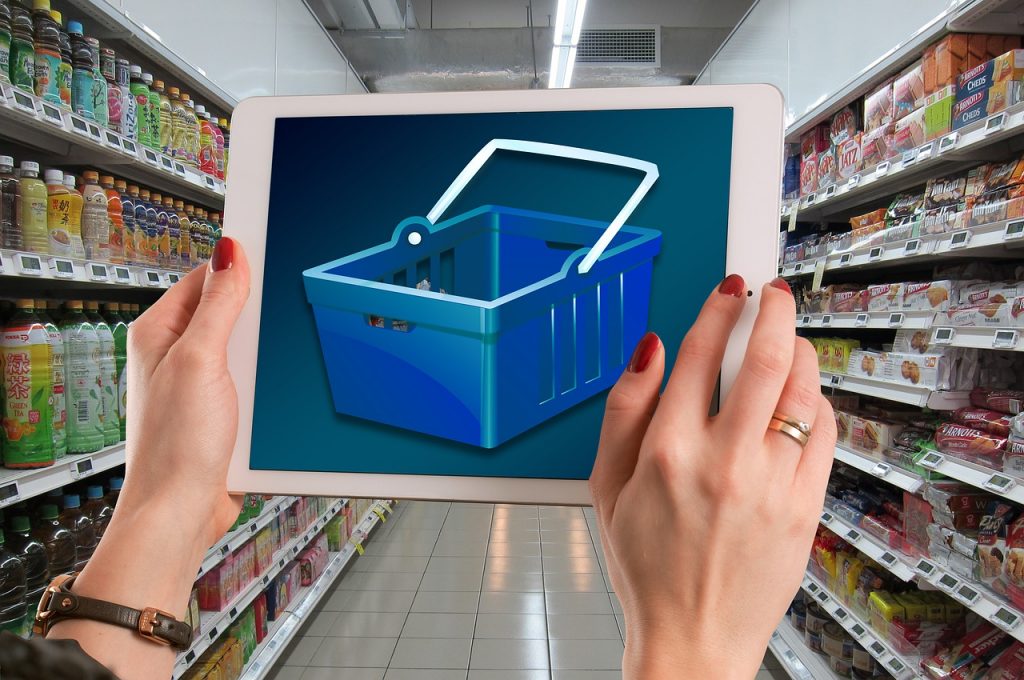 Mãos segurando um tablet com carrinho de compras na tela diante de um corredor de mercearia