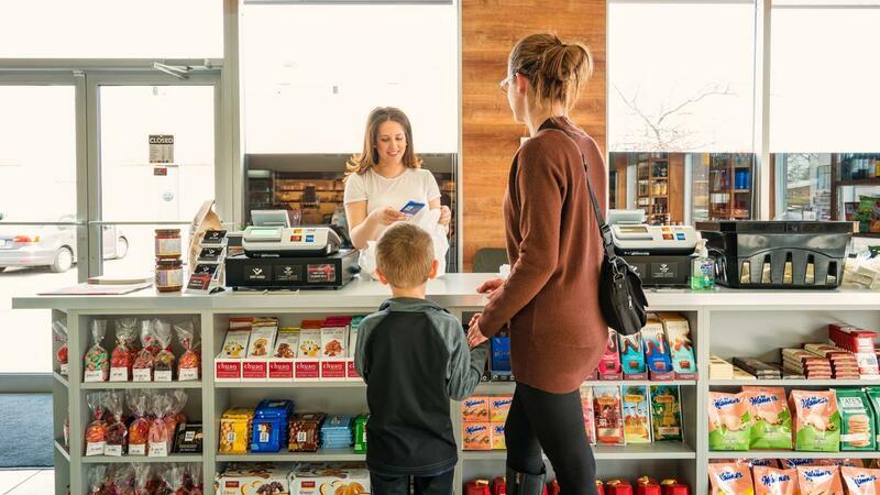 Mulher segurando criança pela mão no checkout da loja, balcão de checkout com produtos variados nas prateleiras e atendente do outro lado