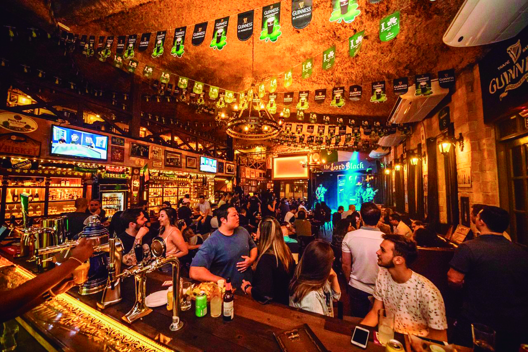 A imagem mostra um bar com decoração rústica, bem iluminado e com pessoas conversando e tomando cerveja.