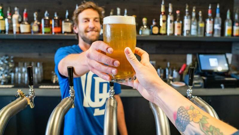 Um homem barista entrega copo de cerveja para pessoa com braço estendido, ao fundo há diversos outros tipos de bebidas
