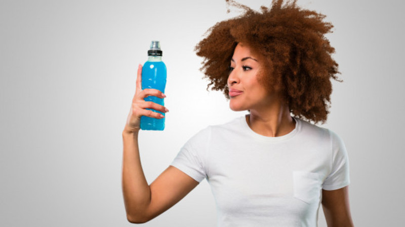 Mulher de camiseta branca, com fundo branco, olhando para uma garrafa de plástico de isotônico com cor azul