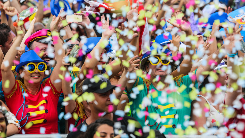 Em um bloco de carnaval, há confetes caindo sobre as pessoas. Elas estão fantasiadas usando óculos e chapéus coloridos.