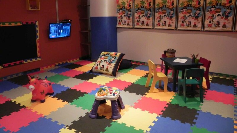  A imagem mostra uma sala infantil com tapete emborrachado colorido, com brinquedos infantis e mesa para colorir. 