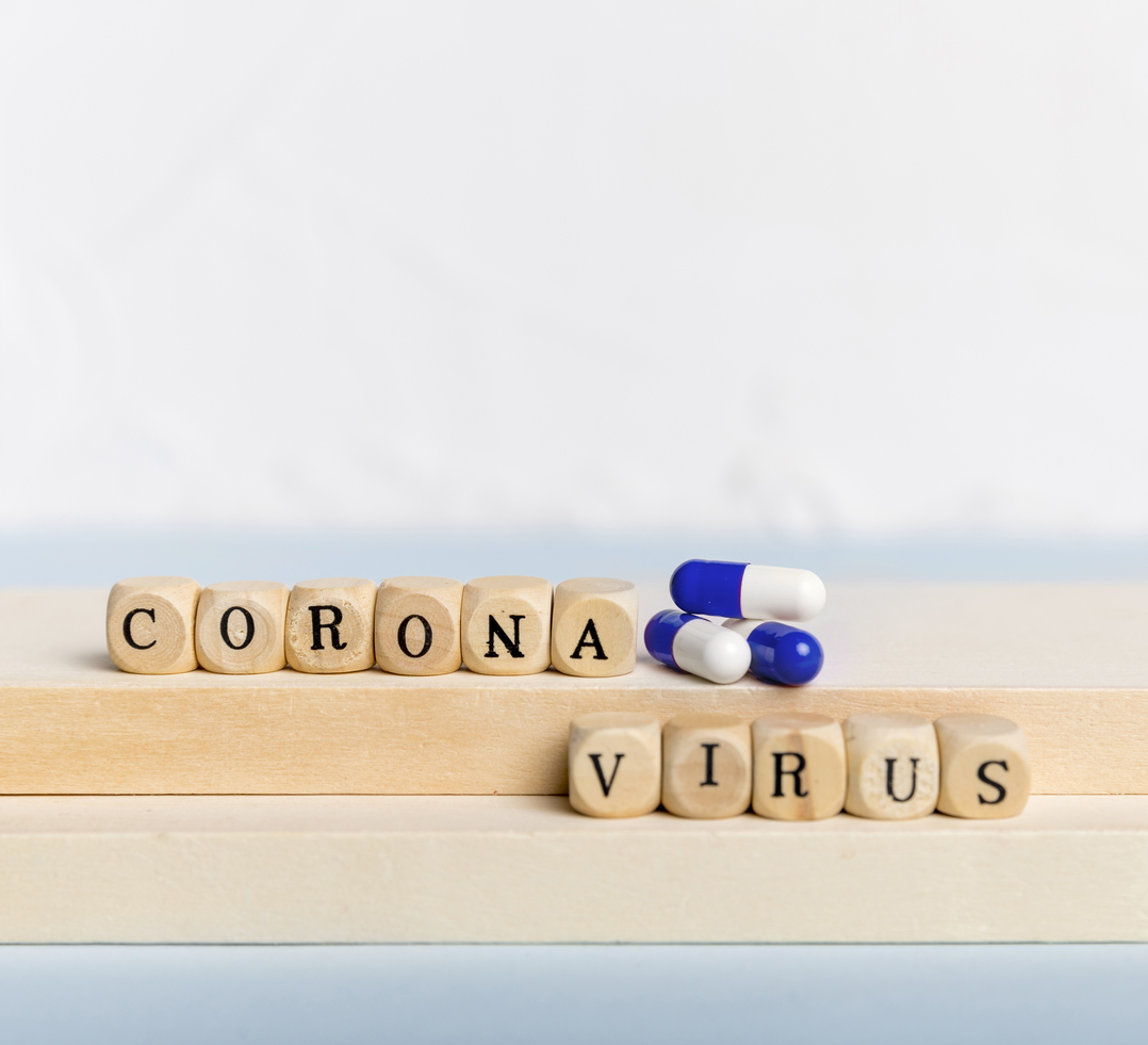 Pecinhas em madeira, cada uma com uma letra, formam a palavra coronavírus. Há também três comprimidos de remédios coloridos.