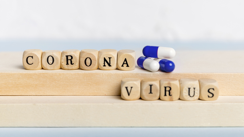 Pecinhas em madeira, cada uma com uma letra, formam a palavra coronavírus. Há também três comprimidos de remédios coloridos.