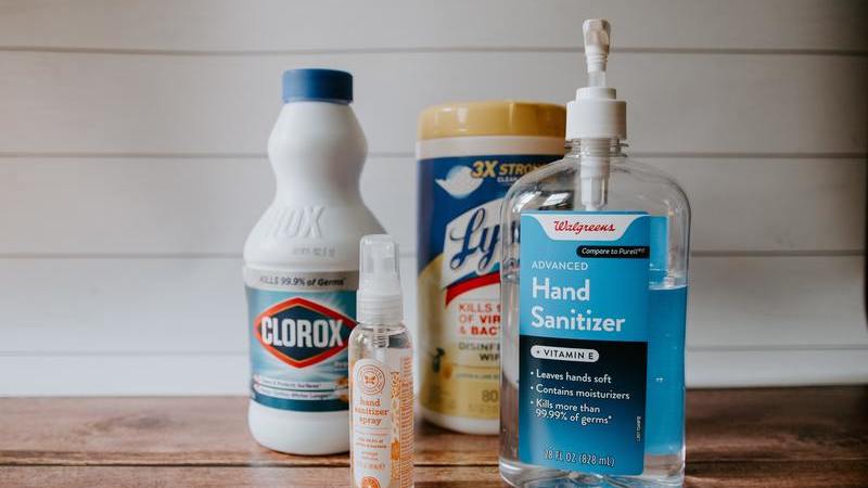 Quatro produtos de limpeza: cloro, álcool em gel, lenço de limpeza e álcool em spray dispostos em uma superfície de madeira.