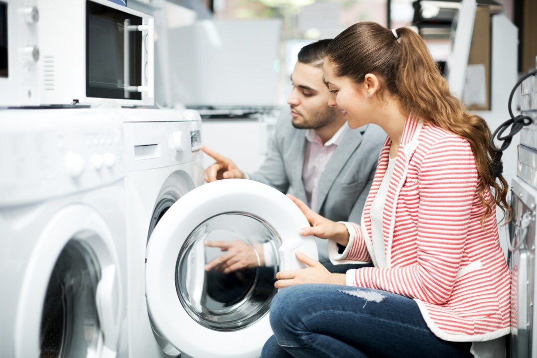 A imagem apresenta um homem e uma mulher ajoelhados e manuseando máquina de lavar com a tampa frontal aberta.