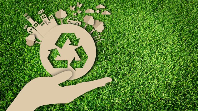 Símbolo da sustentabilidade ilustrada sob a mão de uma pessoa segurando o mundo. Montagem feita sob um fundo de grama verde.
