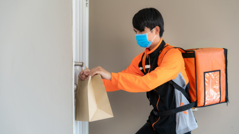 Homem de máscara de proteção segurando sacola de mercadorias. Ele coloca o produto pendurado na porta do cliente.