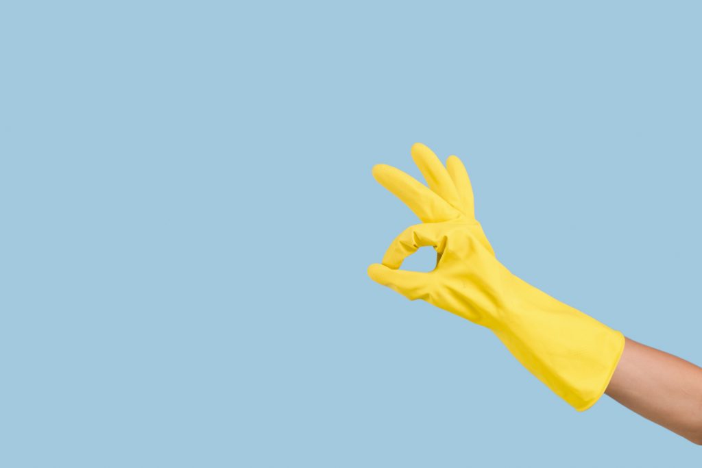 Em um fundo azul, uma mão com luva amarela fazendo o sinal de O.K.