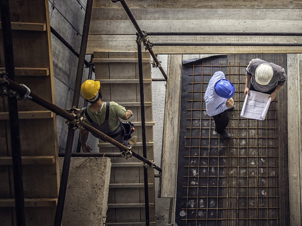 Vista superior de uma construção com três homens trabalhando. Um desce a escada, os outros conversam sobre a planta baixa.