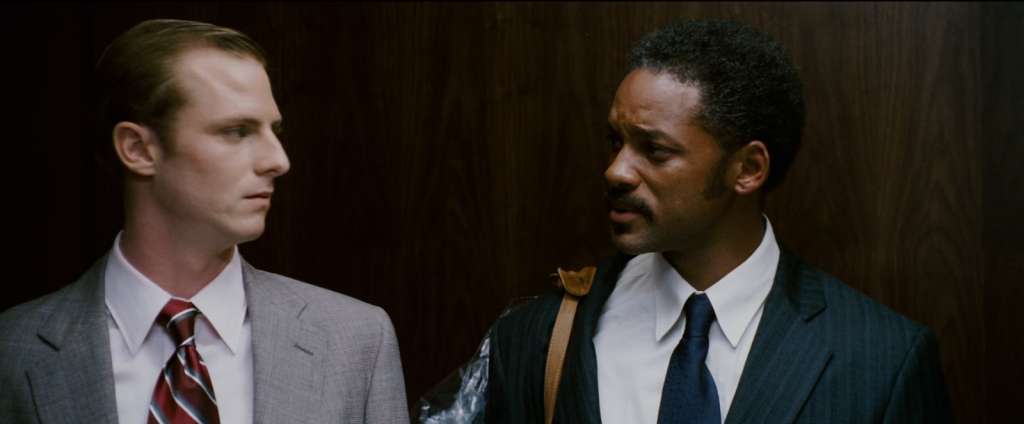 Dois homens de terno se olhando dentro de elevador.