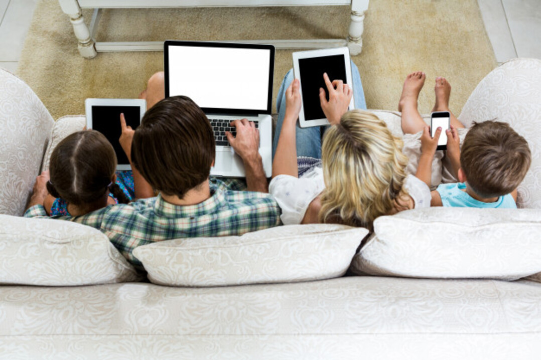 Família sentada no sofá com aparelhos eletrônicos nas mãos: tablet, smartphone e notebook.