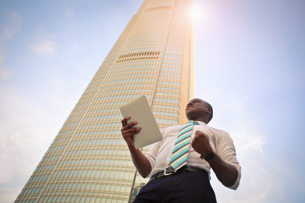 Homem segurando tablet de pé ao lado de um prédio alto. Ele faz gesto de vitória com a mão
