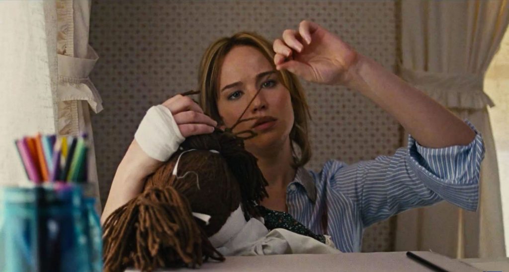 Atriz Jennifer Lawrence com a mão enfaixada puxando um fio de lã da cabeça de uma boneca.