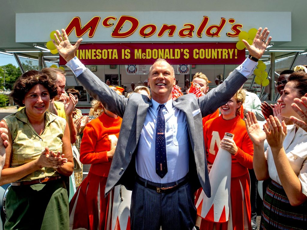 Homem de terno com os braços pra cima cercado de mulheres sorridentes em frente à fachada de uma filial do McDonald’s.