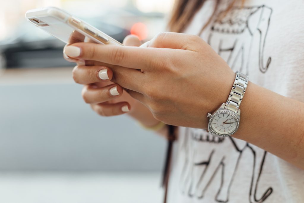 Mão feminina com as unhas pintadas de branco e relógio no pulso segurando smartphone.