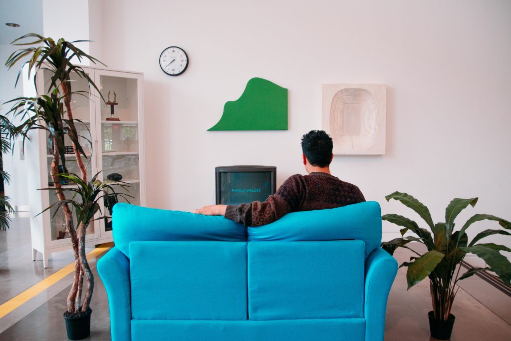 Em uma sala, homem está de costas para a foto. Ele está sentado em um sofá azul olhando para a televisão.