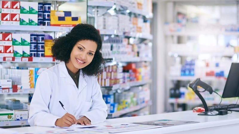 Mulher de jaleco branco sorrindo apoiada sobre balcão de farmácia segurando caneta sobre papel.