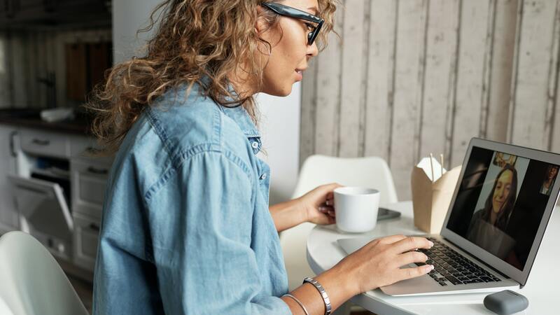 Mulher de óculos e blusa jeans sentada em uma cadeira branca. Ela faz uma vídeo chamada com outra mulher usando notebook. 