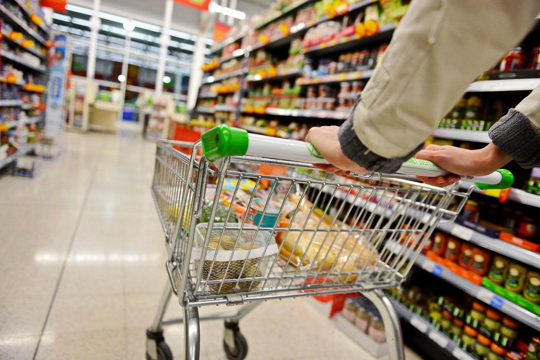 Braços de pessoa empurrando carrinho de compras em corredor de supermercado cercado de duas prateleiras de alimentos.