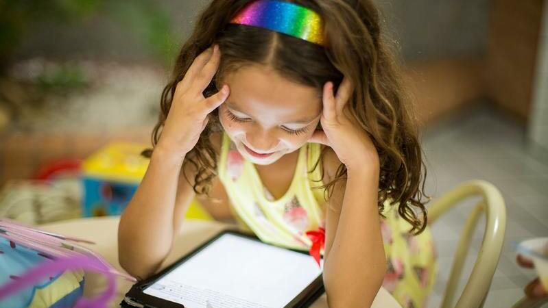Menina olhando tablet com as mãos na cabeça. Ela está sentada em uma mesa e usa uma tiara colorida.