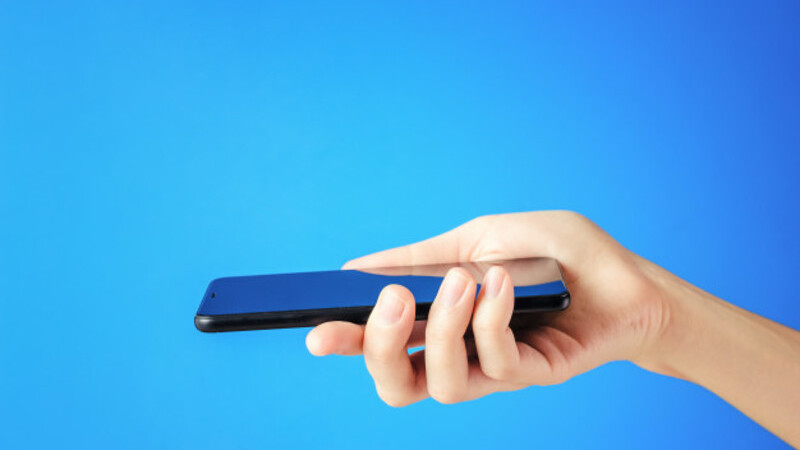 Em um fundo azul uma mão segura um smartphone preto. 