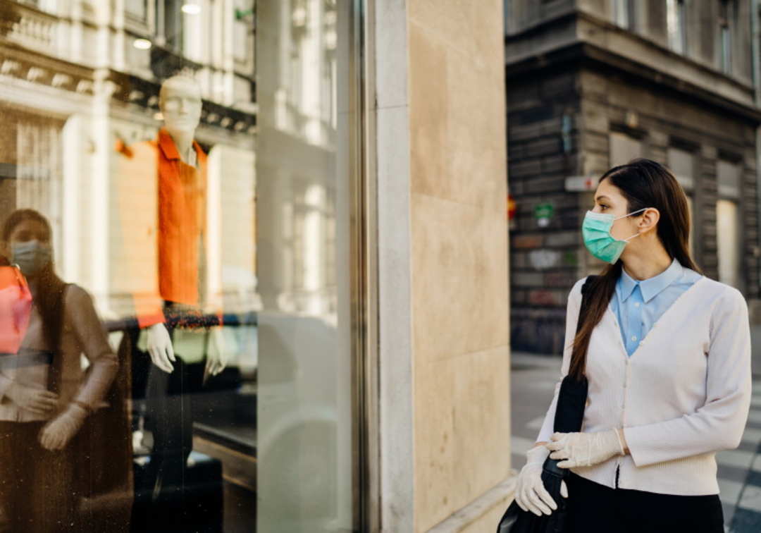 Mulher usando luvas e máscara facial de proteção andando na calçada e olhando para vitrine à sua esquerda.