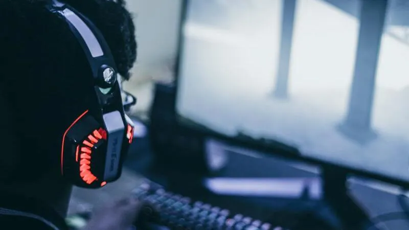 Homem de costas usando headset gamer no escuro em frente a monitor com jogo na tela.
